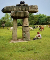 a woman sits on a grassy field next to a large rock